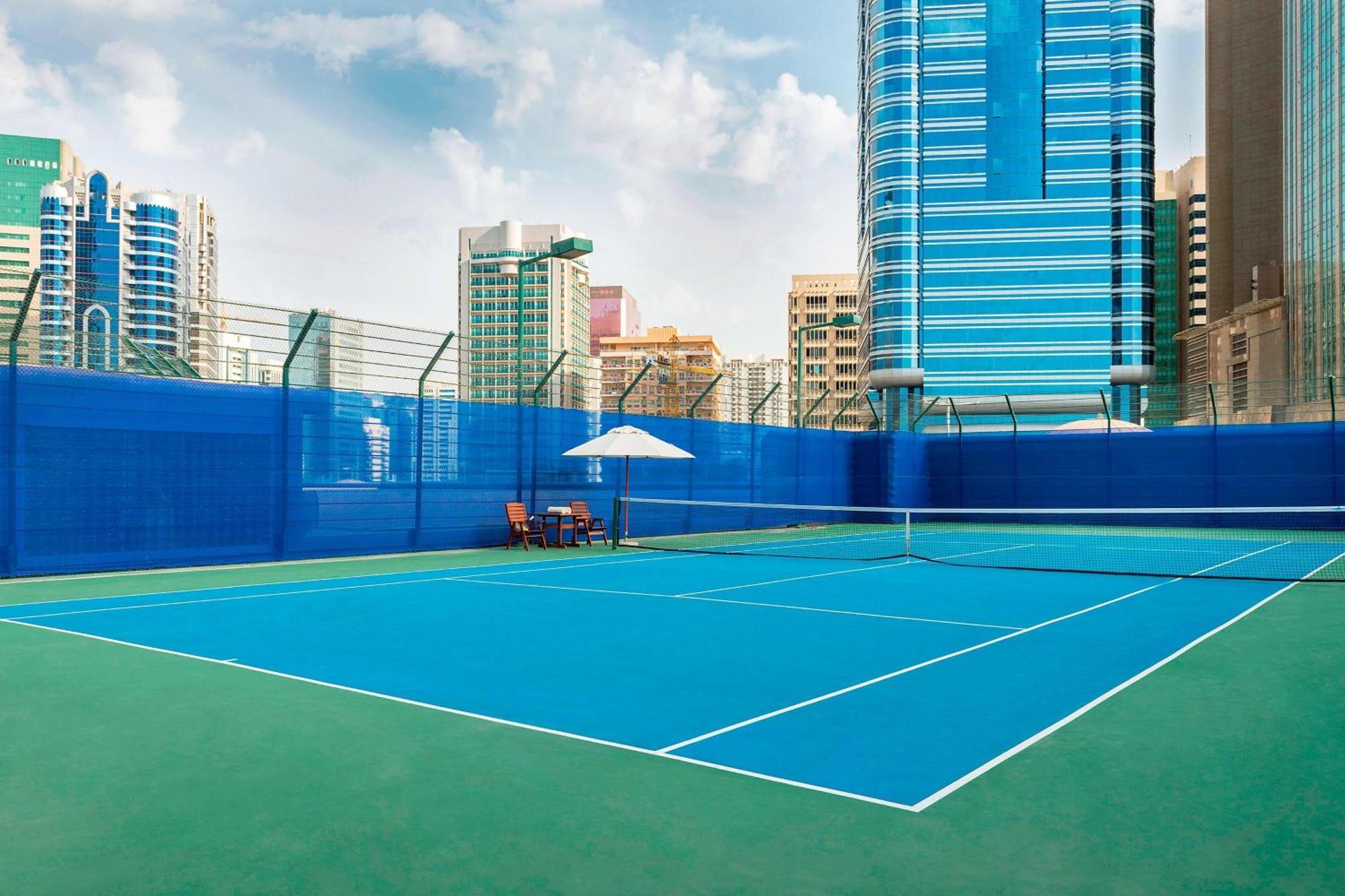 Sheraton Abu Dhabi Hotel & Resort Exterior photo The photo shows a tennis court situated in an urban environment with tall buildings in the background. The court features a vibrant blue playing surface, surrounded by a green area. There is a net in the center, and a couple of chairs and an umbrella